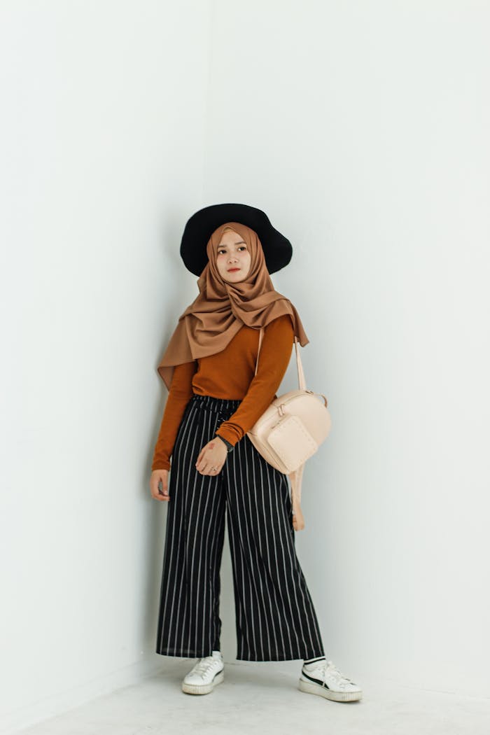 Fashionably dressed woman in hijab posing against a minimalist white wall, showcasing a stylish look.